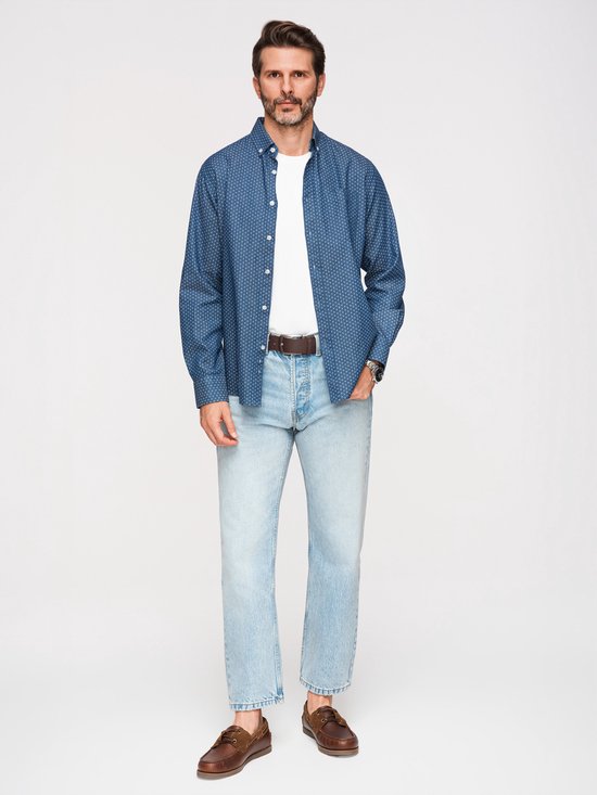 Man met baard staat, draagt een blauw overhemd met witte stipjes, wit t-shirt, spijkerbroek, bruine riem en bruine schoenen.