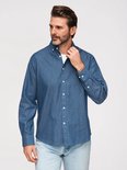 Man met baard, draagt een blauw overhemd met witte stipjes, wit t-shirt en spijkerbroek, raakt de kraag aan.
