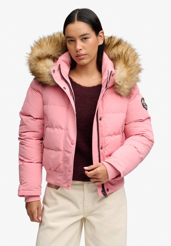 Vrouw in roze Superdry bomberjack met bontkraag en beige broek.