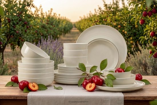 Een witte tafel met daarop een uitgebreide set beige serviesgoed, versierd met pruimen en groene bladeren, met een pruimenboomgaard op de achtergrond.