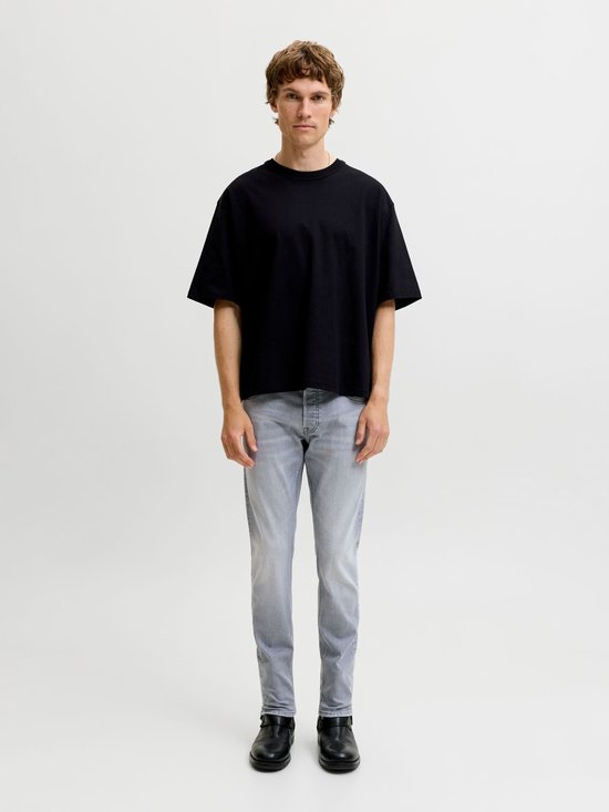 Man in zwarte oversized t-shirt en grijze jeans met rechte pijpen, staand in de studio.