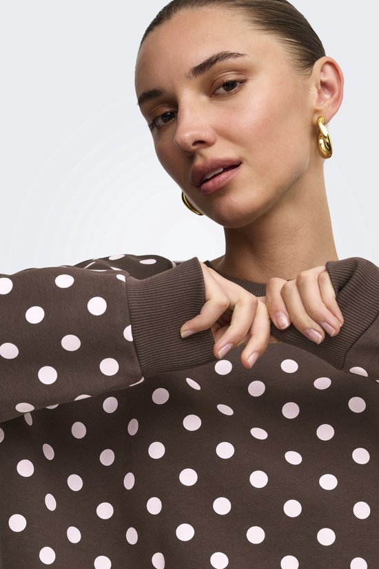 Close-up van een bruine sweater met roze polkadots, gedragen door een model met gouden oorbellen.