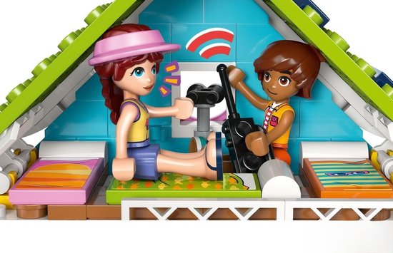 Twee vriendinnen in een LEGO huisje, één met roze pet, zittend op een groen kleed met op de achtergrond een raam met zend-signalen.