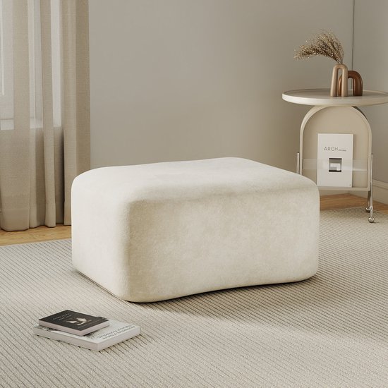 Beige fluwelen poef met zachte, afgeronde vorm op een gestreept tapijt in een lichte woonkamer.