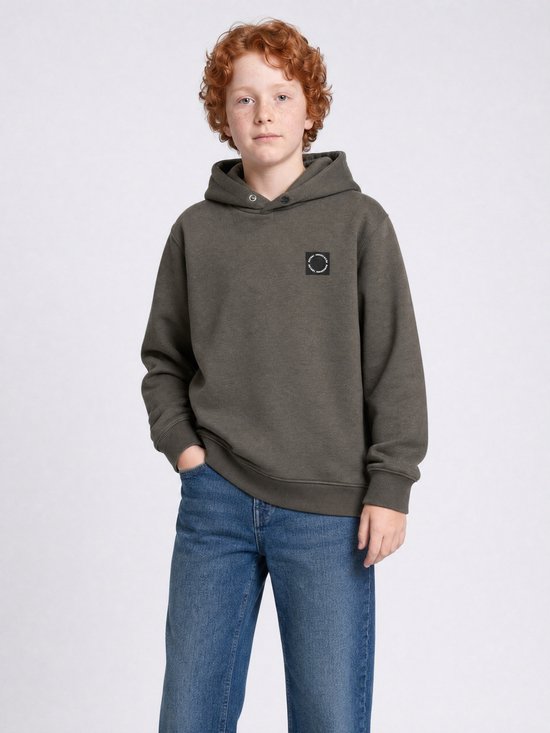 Jongen met krullend rood haar, met handen in de zakken, in een donkergroene hoodie en blauwe jeans.