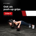 Vrouw met zwarte halterset in push-up houding, rode handgrepen.