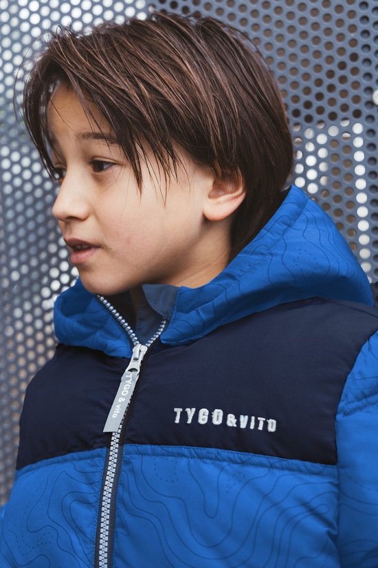 Close-up van een jongen in een blauw gewatteerd jack met zwart borstpaneel en contourpatroon. Rits met TYGO & VITO tekst.