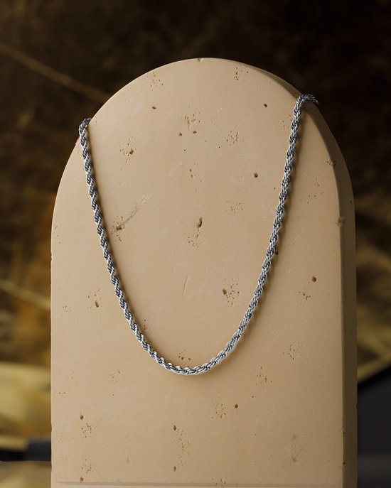 Een zilverkleurige, gedraaide touwketting tentoongesteld op een beige, afgeronde displaystandaard tegen een warme, gebogen achtergrond.