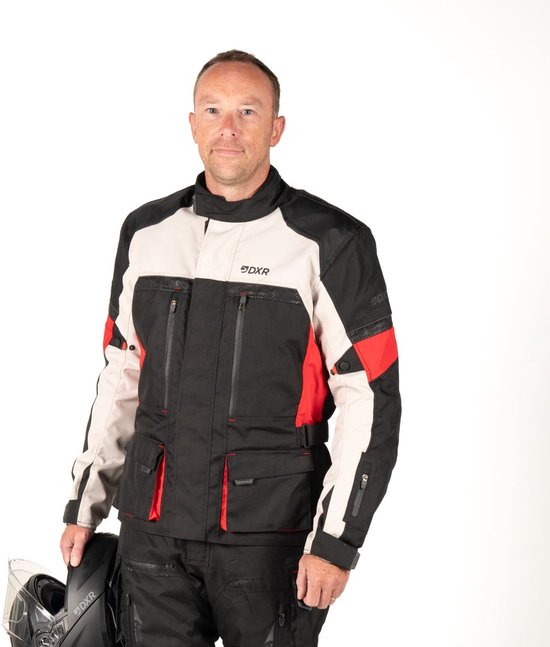 Man draagt een zwarte motorjas met lichtgrijze en rode accenten, en zwarte motorbroek. Houdt een helm vast.