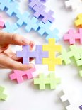 Hand houdt twee Plus-Plus bouwstenen vast, paars en geel, om ze te verbinden. Andere pastelkleurige stenen liggen verspreid.