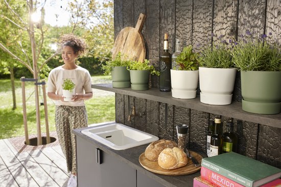 Vrouw met plant in witte pot, bij een buitenkeuken met planten en broodjes.