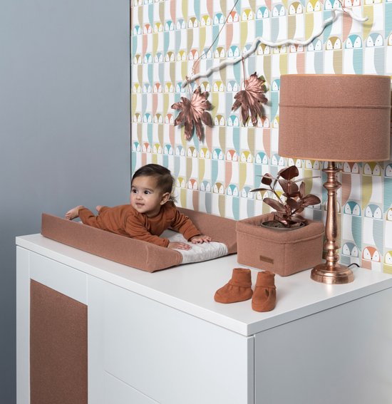 Bébé en pyjama marron est allongé sur une commode avec un matelas à langer brun rouille et des paniers assortis.