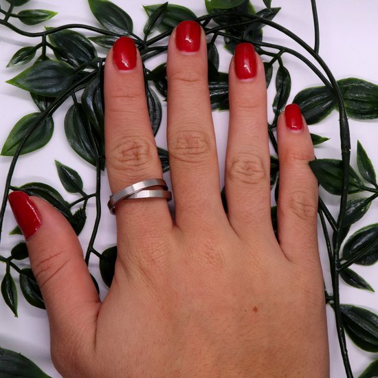 Hand met rode nagellak en een zilverkleurige ring om de middelvinger, omringd door groene bladeren.
