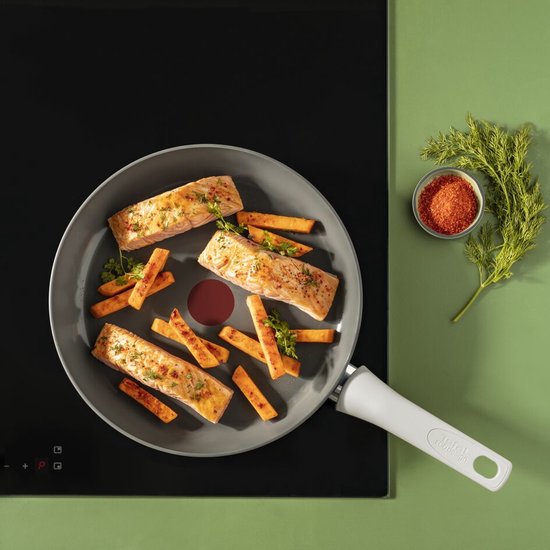 Grijze koekenpan met drie stukken zalm, frietjes, peterselie en dille op een zwarte inductiekookplaat.