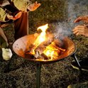 Vuurschaal met brandend hout, buiten op gras, met mensen die hun handen warmen.