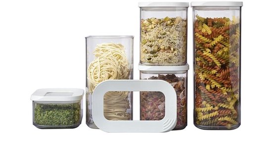 Six boîtes de conservation transparentes avec couvercles blancs, remplies de pâtes, d'herbes, de muesli et de fruits secs.