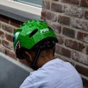 Lichtgroene kinderfietshelm met dinosaurusprint, grote blauwe ogen en scherpe tanden. Draagt een jongen.