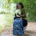 Vrouw met donker haar en groene trui zit op een bosbodem naast een blauwe rugzak met geometrisch patroon.