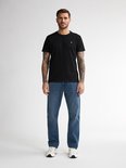 Man met tatoeages draagt een zwarte t-shirt en blauwe jeans.