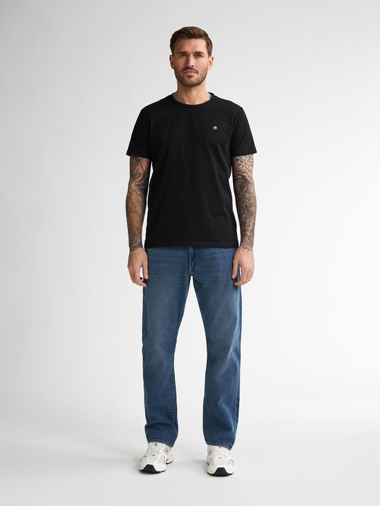 Man met tatoeages draagt een zwarte t-shirt en blauwe jeans.