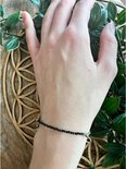 Zwarte toermalijn kralenarmband met zilverkleurig hartje, gedragen om een pols, naast groene bladeren.