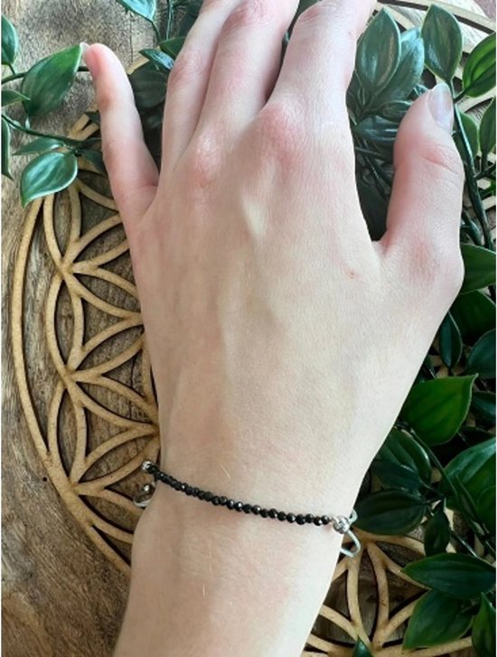 Zwarte toermalijn kralenarmband met zilverkleurig hartje, gedragen om een pols, naast groene bladeren.