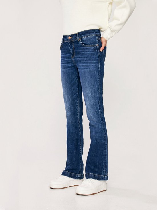 Model in een witte trui en donkerblauwe flared jeans, gefotografeerd vanaf de zijkant.