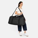Model draagt een zwarte Eastpak reistas over de schouder, gecombineerd met een wit T-shirt en donkerblauwe broek.