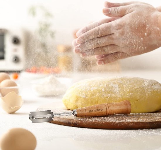 Bakkers handen strooien bloem over een deeg met een deegmes ernaast op een houten plank.