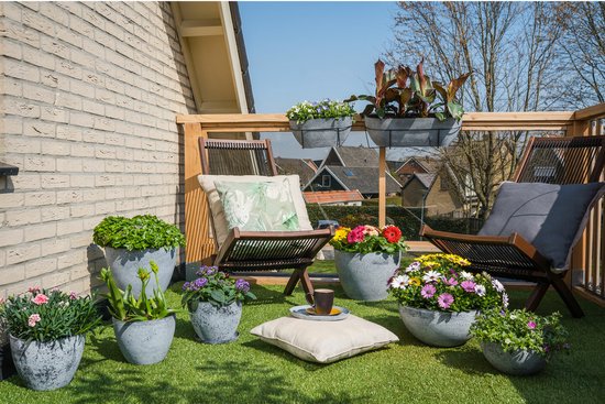 Grijze betonnen plantenbakken met kleurrijke bloemen en groene planten op een balkon met tuinmeubilair.