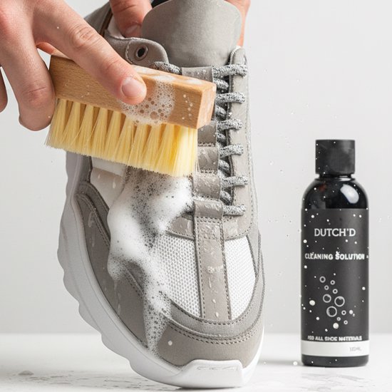 Een grijze sneaker wordt schoongemaakt met een gele borstel en schuim. Naast de schoen staat een flesje "DUTCH'D CLEANING SOLUTION".