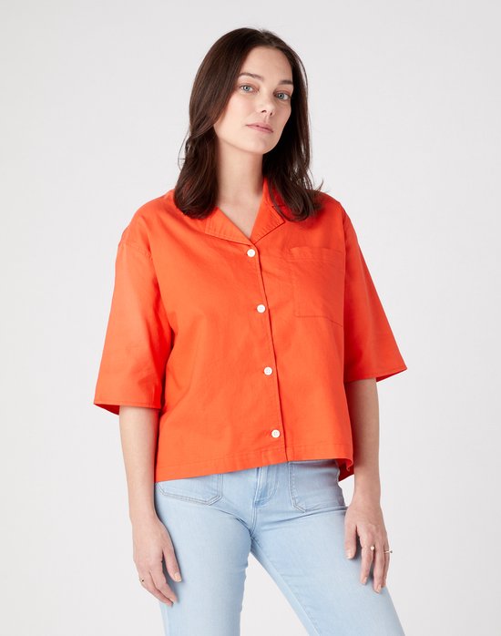 Model met een oranje blouse en lichte jeans, kijkt recht vooruit.