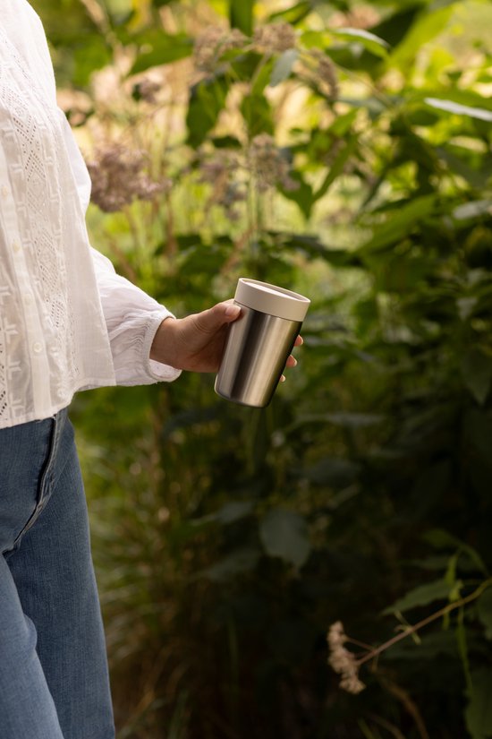 Vrouw in witte blouse houdt een zilverkleurige thermosbeker vast in de tuin.