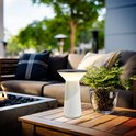 Witte Pilia tafellamp op een houten tafel naast een plant en een bank met open haard op een terras.