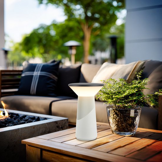Witte Pilia tafellamp op een houten tafel naast een plant en een bank met open haard op een terras.