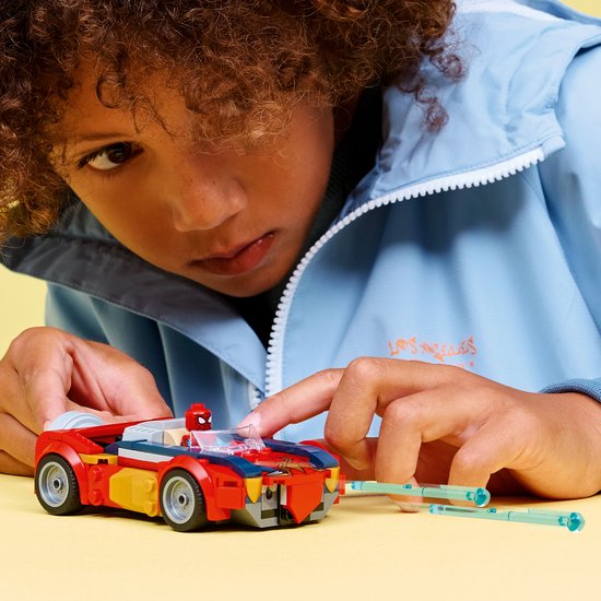 Kind met krullend haar bouwt rode raceauto met Spiderman-figuur en blauwe jas.