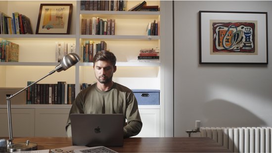 Man zit op een bureaustoel aan een houten bureau met een laptop, een radiator staat rechts.