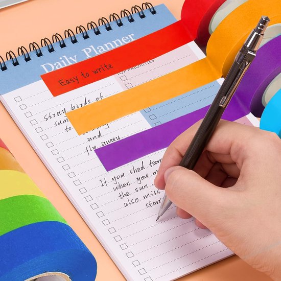 Een hand schrijft op een dagplanner. Er zijn gekleurde stroken tape gebruikt om het plan te markeren. Verschillende rollen gekleurde tape liggen ernaast.