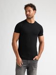 Man met handen in broekzakken, draagt zwart T-shirt met korte mouwen en grijze jeans.