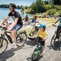 Man en kinderen fietsen langs een meer, met een fietskar achter de fietsen.
