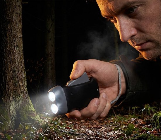 Man met donkere jas houdt een zwarte knijpkat dynamo zaklamp met twee witte lichten die schijnen op een boom.