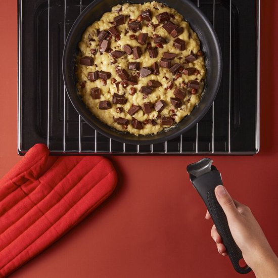 Chocoladekoekjesdeeg met chocoladestukjes in een zwarte pan op een ovenrooster, met rode ovenwanten en een afneembare handgreep.