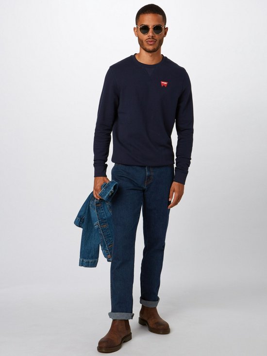 Man met zonnebril in donkerblauwe Wrangler jeans, marineblauwe trui met rood logo, bruine laarzen.