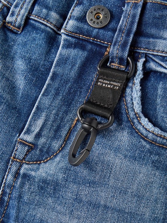 Rits en knoop van een spijkerbroek met een zwarte stoffen lus met karabijnhaak en "ORG.DENIM PRODUCTS BY NAME IT" tekst.