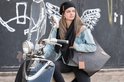 Jonge vrouw met zwarte gebreide muts en sjaal, op een scooter, met een tas.