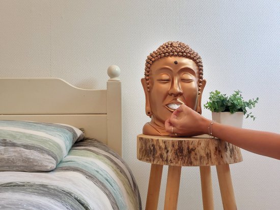 Gouden Boeddha hoofd met witte tissue uit de mond, op een houten bijzettafel naast een bed.