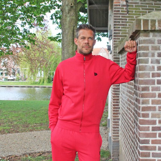 Man met rood trainingsvest en broek, leunend tegen een bakstenen muur in een park met water.