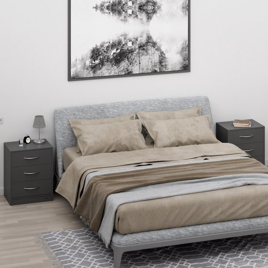 Grijs nachtkastje met 3 lades naast een bed met beige beddengoed. Een lampje en wekker staan op het nachtkastje.