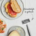 Râpe à fromage à côté d'un sandwich grillé ouvert avec du fromage et de la tomate et une assiette de fromage râpé.