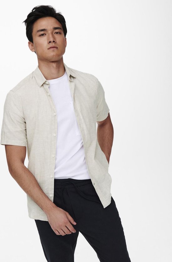 Man met een beige linnen mix overhemd met korte mouwen en een witte t-shirt, met handen in zakken.
